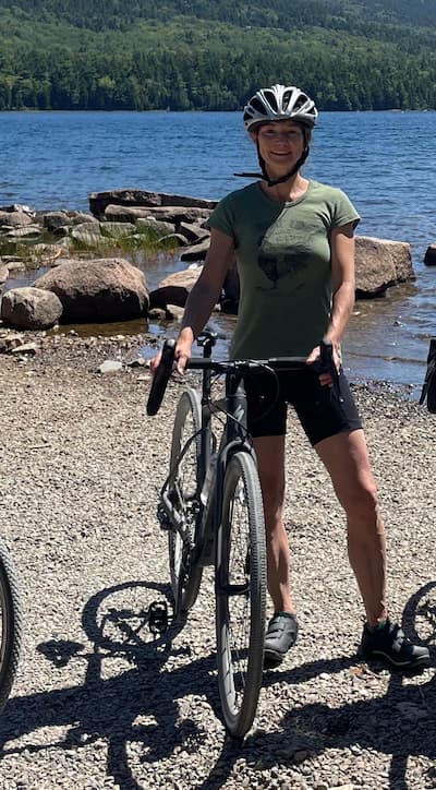 Holly Conn standing with her bicycle in front of a lake in summer.