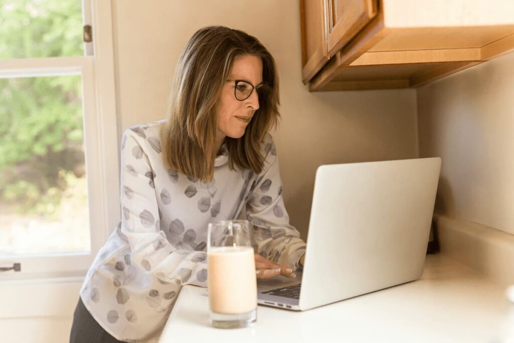 Woman using laptop at kitchen counter with glass of milk in foreground.  Photo by LinkedIn Sales Solutions on Unsplash