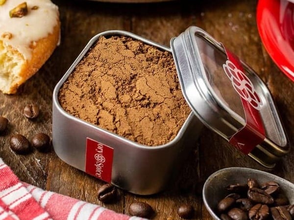 A tin of spiced drink mix by Savory Spice.   A spoonful of unground coffee beans and an iced cake surround the tin.