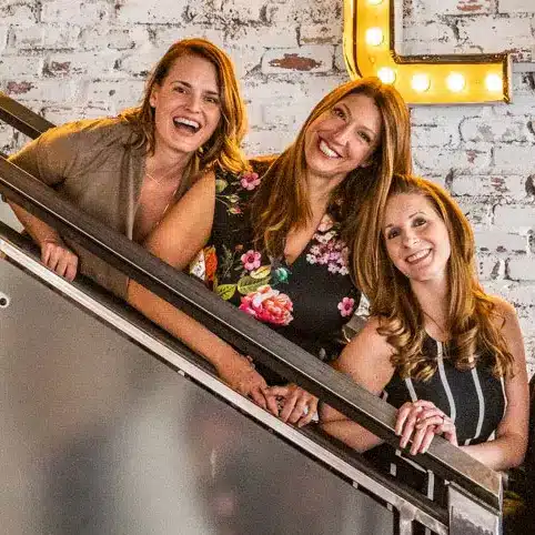 Three smiling women standing on a staircase, looking directly at the camera.